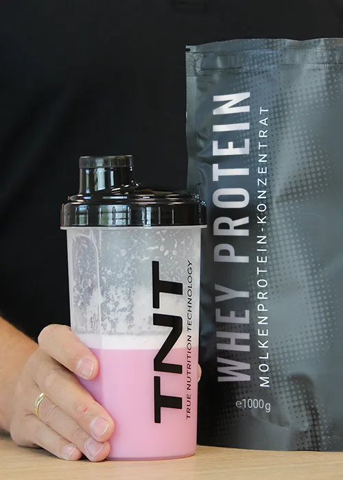 Ein Beutel TNT Whey Protein Konzentrat mit einem gefüllten Shaker im Vordergrund